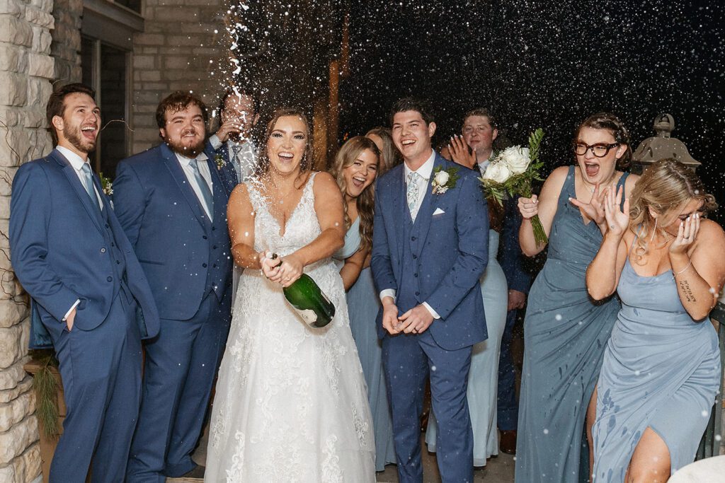Bride doing a champagne pop with the groom and wedding party having fun in the background