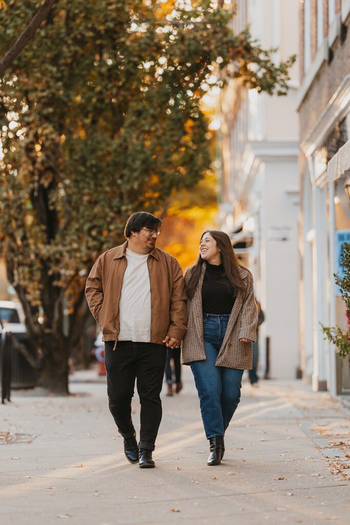 A cozy, romantic photo of the couple, Alexa and Joe, strolling hand-in-hand through the charming streets of downtown Palmer Square, adorned with fall decorations.