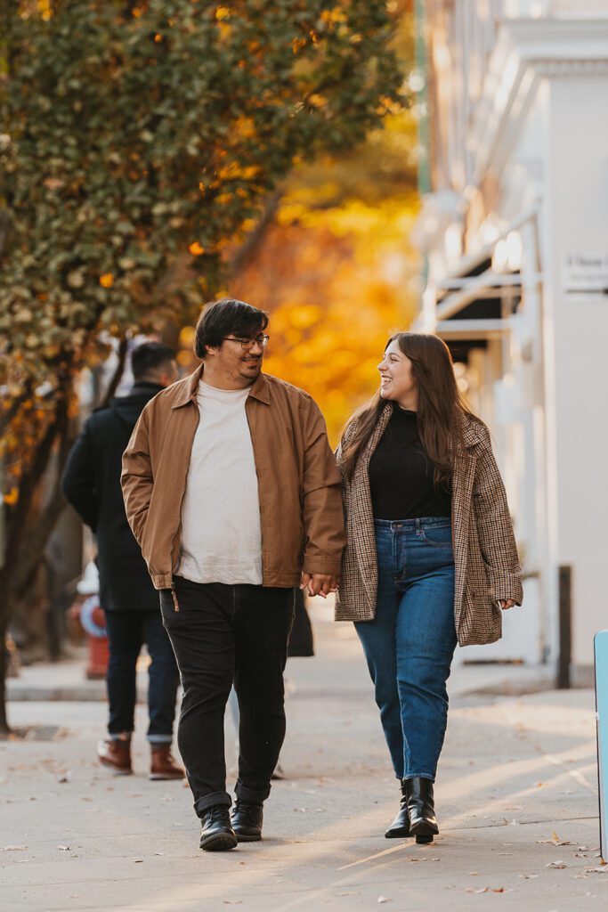 A cozy, romantic photo of the couple, Alexa and Joe, strolling hand-in-hand through the charming streets of downtown Palmer Square, adorned with fall decorations.