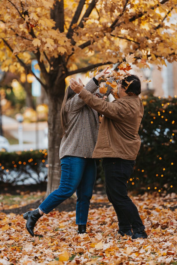 Candid laughter and joy radiate from Alexa and Joe as they indulge in a playful leaf fight, a moment of pure bliss during their fall engagement photo shoot in Palmer Square