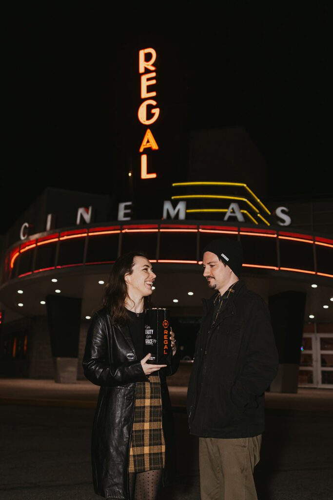 Engaged couple, Christine and Joe, striking a casual pose with the charming facade of a movie theater in the background, perfect for their movie theater-themed engagement photos.