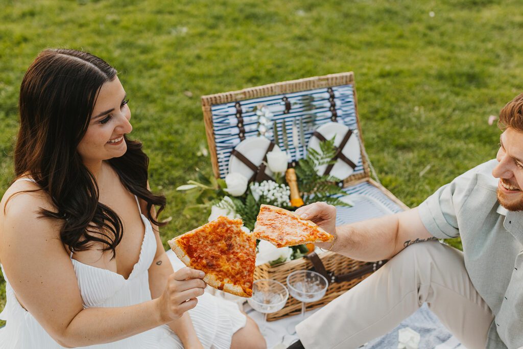 a sweet couple feeding each other pizza in a park