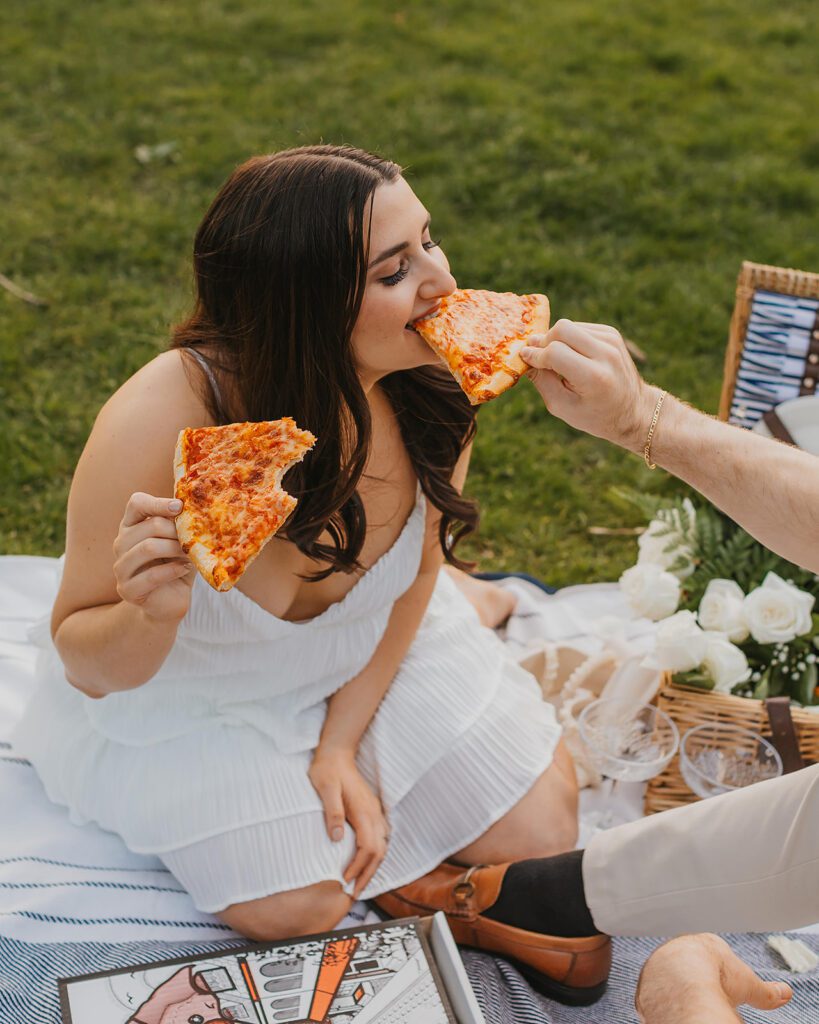 a sweet couple feeding each other pizza in a park