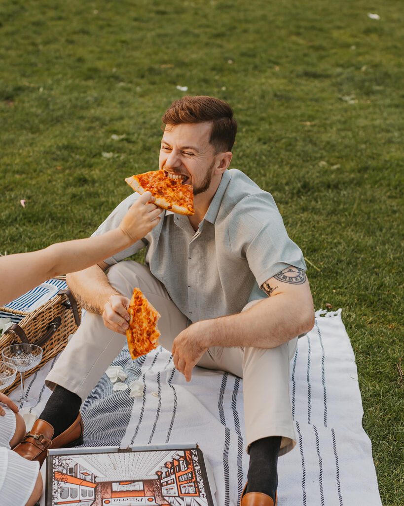 a sweet couple feeding each other pizza in a park