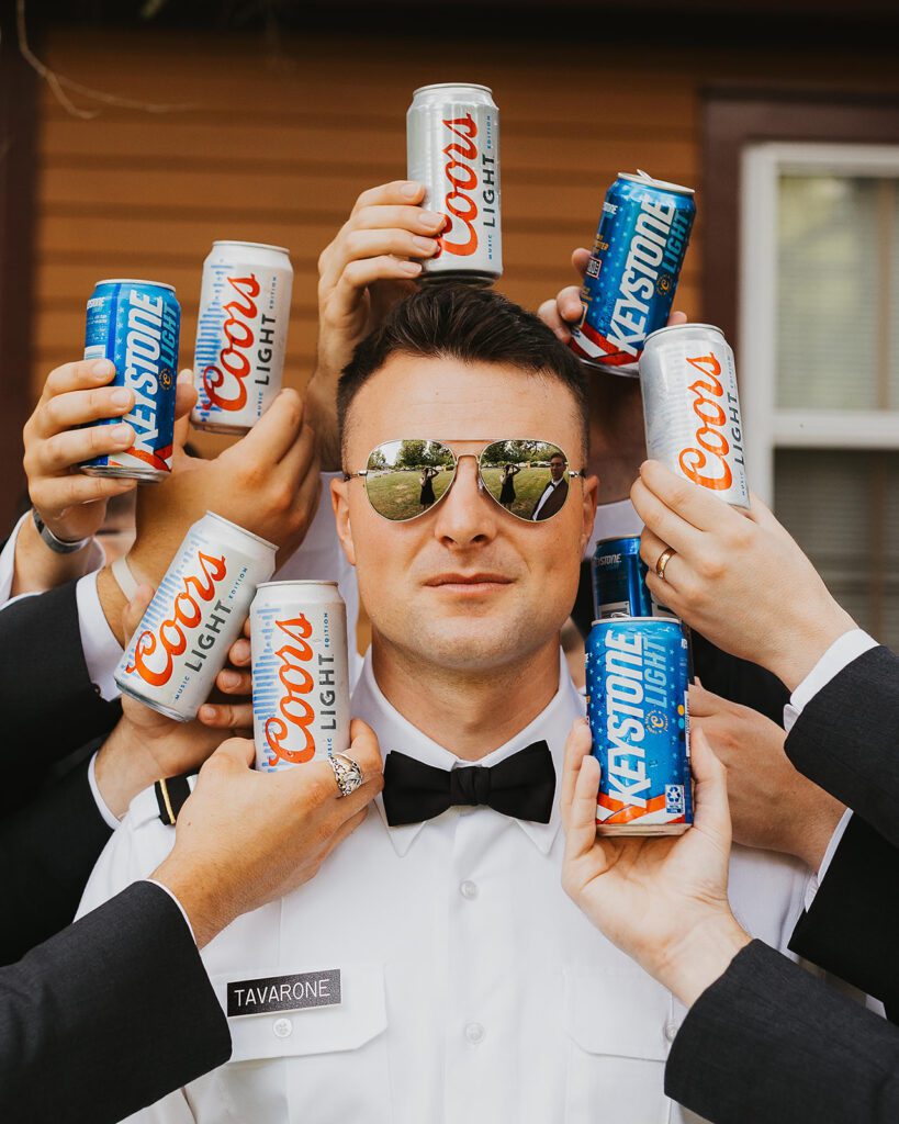 The groom, wearing aviator sunglasses and a crisp white shirt with a bow tie, poses confidently as his groomsmen hold cans of Coors Light and Keystone Light around him