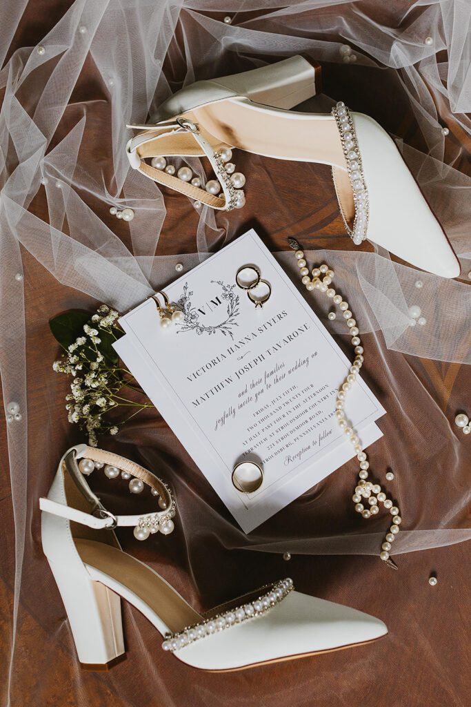 A close-up shot of the bride's pearl-embellished wedding shoes, accompanied by a velvet ring box displaying both the wedding bands and the engagement ring