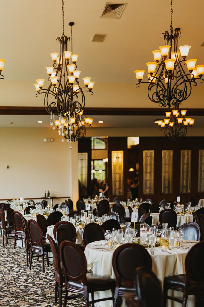 A warmly lit reception room with chandeliers hanging from the ceiling. Tables are elegantly set with white tablecloths, glassware, and floral centerpieces