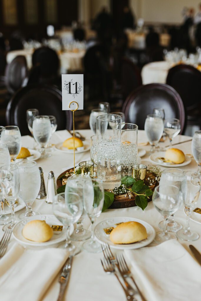 A warmly lit reception room with chandeliers hanging from the ceiling. Tables are elegantly set with white tablecloths, glassware, and floral centerpieces