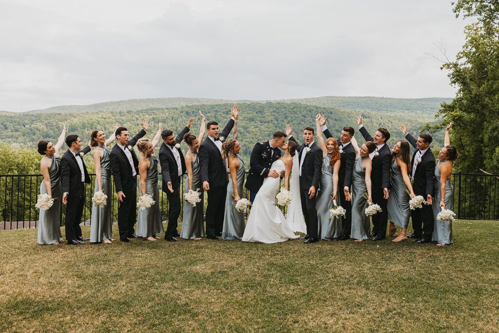 The entire wedding party celebrates together, with bridesmaids and groomsmen raising their arms in cheer as the bride and groom share a romantic kiss. The party is positioned on a grassy lawn with a scenic mountain landscape in the background at Stroudsmoor Country Inn