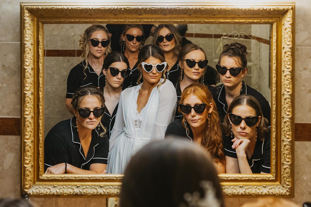 The bride and her bridesmaids, all wearing matching black pajamas pose in front of a large mirror, Each wears heart-shaped sunglasses