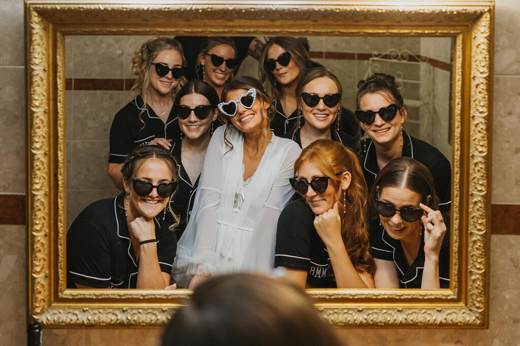 The bride and her bridesmaids, all wearing matching black pajamas pose in front of a large mirror, Each wears heart-shaped sunglasses