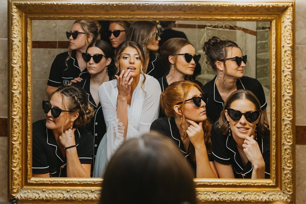 The bride and her bridesmaids, all wearing matching black pajamas pose in front of a large mirror, Each wears heart-shaped sunglasses