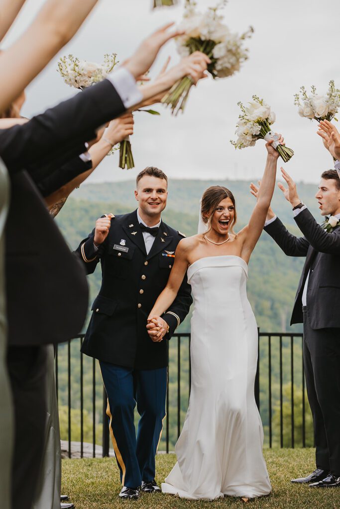 The entire wedding party celebrates together, with bridesmaids and groomsmen raising their arms in cheer as the bride and groom share a romantic kiss. The party is positioned on a grassy lawn with a scenic mountain landscape in the background at Stroudsmoor Country Inn