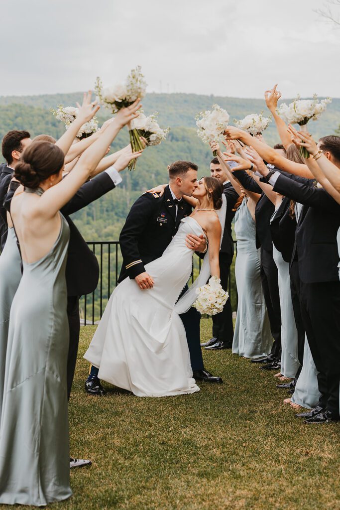 The entire wedding party celebrates together, with bridesmaids and groomsmen raising their arms in cheer as the bride and groom share a romantic kiss. The party is positioned on a grassy lawn with a scenic mountain landscape in the background at Stroudsmoor Country Inn