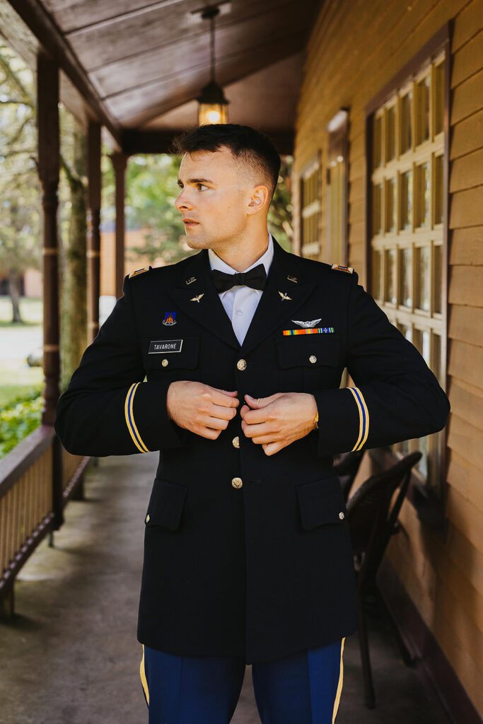 A groom in a formal military uniform adjusts his jacket on a wooden porch