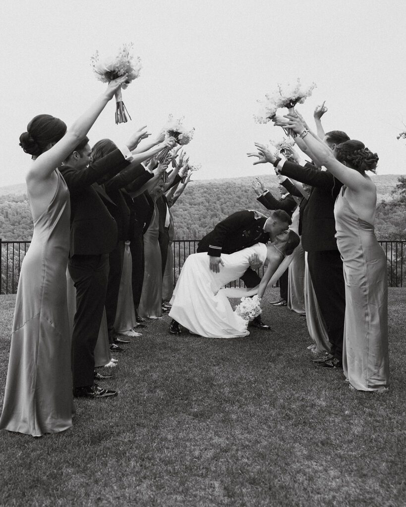 The entire wedding party celebrates together, with bridesmaids and groomsmen raising their arms in cheer as the bride and groom share a romantic kiss. The party is positioned on a grassy lawn with a scenic mountain landscape in the background at Stroudsmoor Country Inn