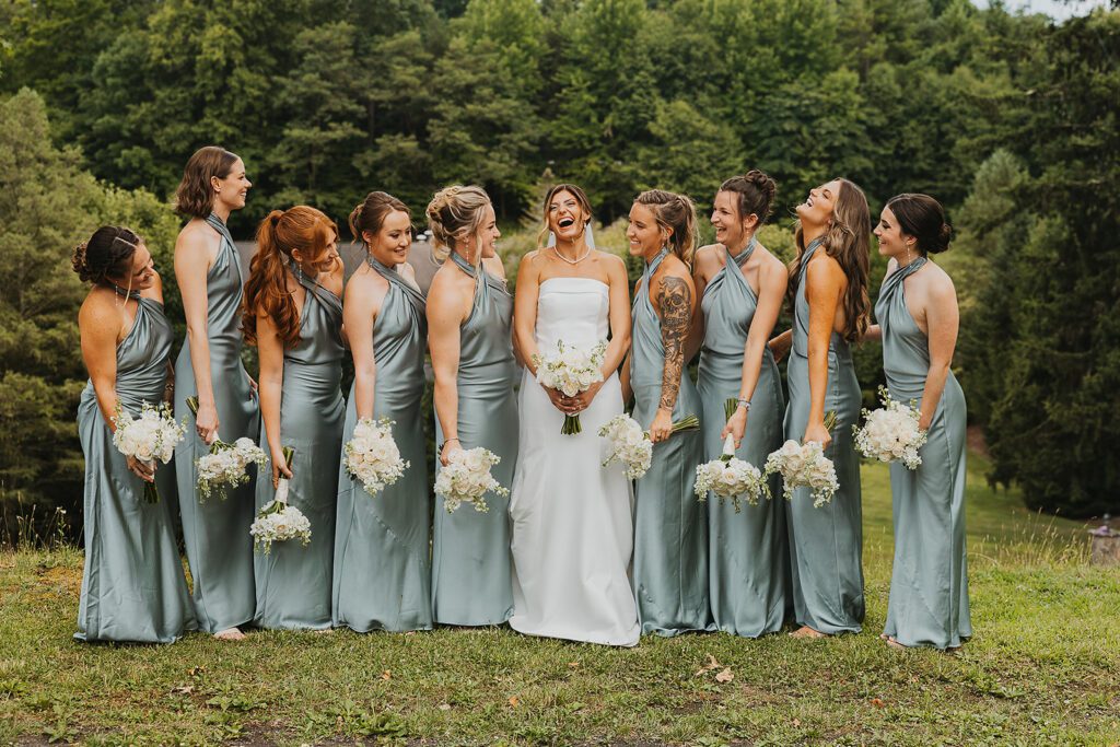 The bride stands joyfully laughing in the center of her bridesmaids, who are all wearing matching satin sage-green gowns