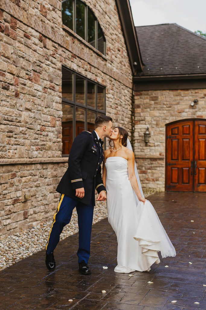 The bride and groom share a quiet moment walking hand-in-hand in front of a beautiful stone mansion. The groom wears his military uniform, and the bride is in a strapless gown