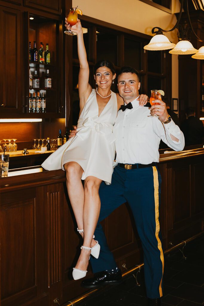 The bride, sitting on the bar, raises her cocktail glass high in celebration while the groom, standing beside her, smiles and holds his own drink